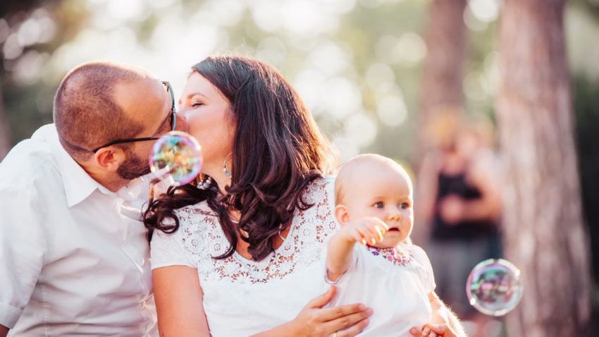 A tender family moment captured with a couple kissing and a baby holding bubbles outdoors.